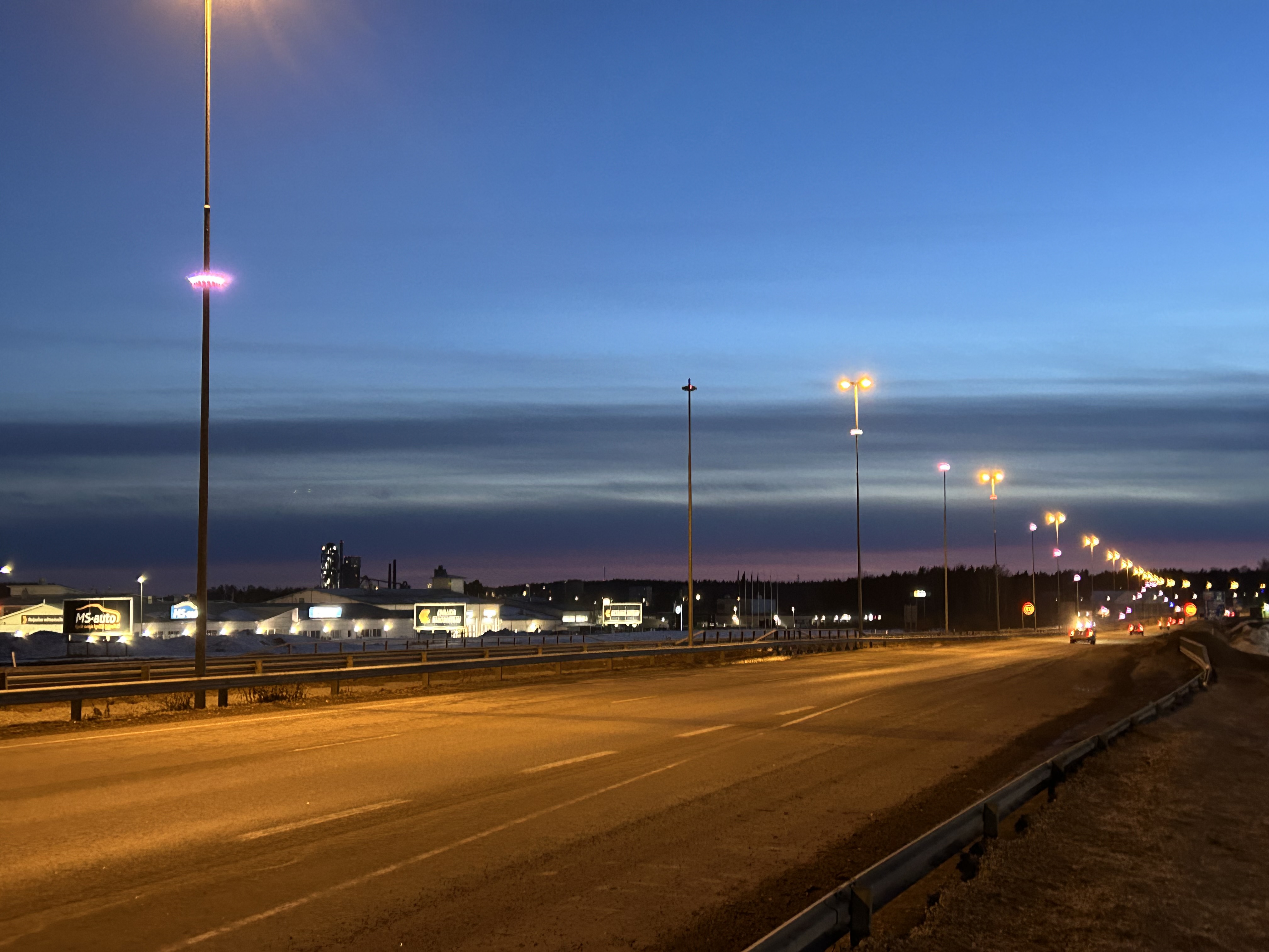 Valtatie 6 pimeällä Lappeenrannan Myllymäen kohdalla, jossa keskikaistan valopylväissä valotaideteosvaloja. Autoja, tienpengertä, rakennuksia.